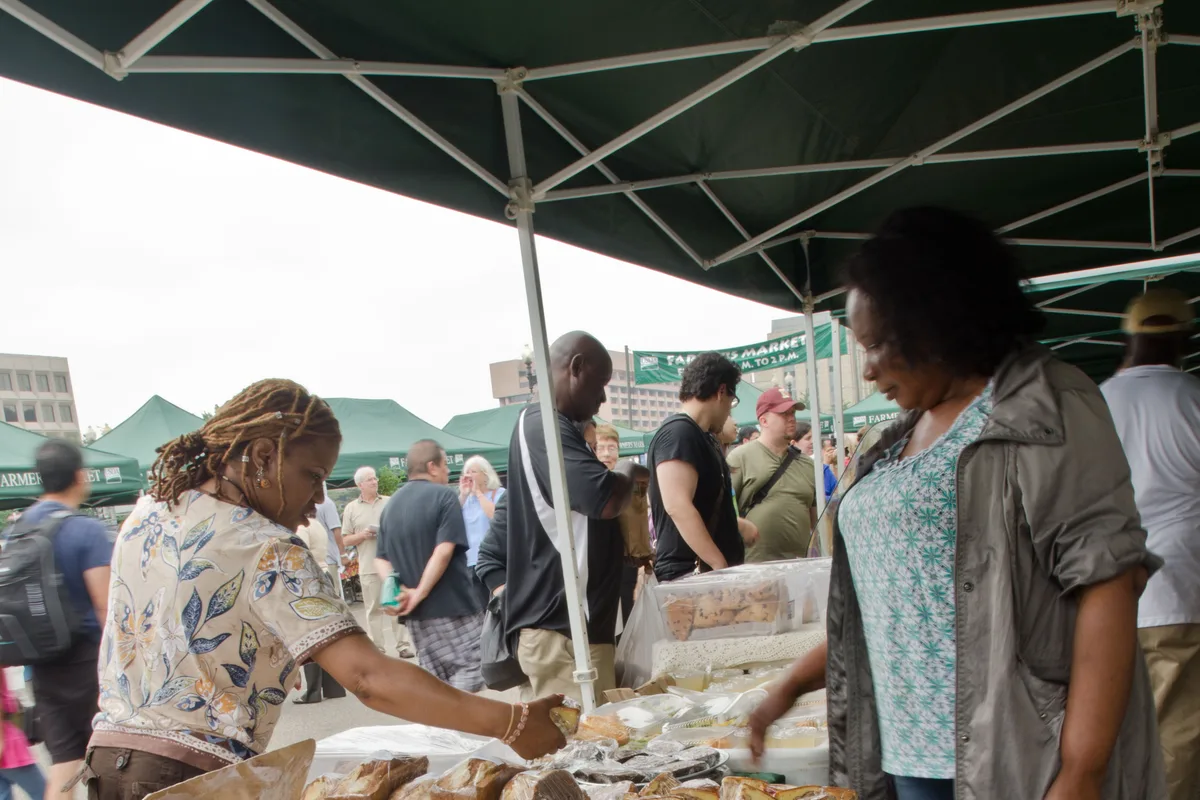 A vendor hands baked goods to a customer at an outdoor farmers market stall.